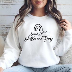Könnte beinhalten: Weißes Sweatshirt mit einem schwarzen Regenbogen-Design und dem Schriftzug "Same Shit Different Day" in Schreibschrift. Ein lässiges Oberteil für den Alltag.