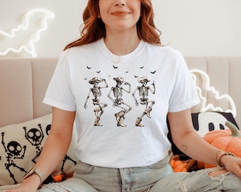 May include: White t-shirt with a graphic of three dancing skeletons in cowboy hats and boots. Small black bat silhouettes are above the skeletons. A fun and festive design for Halloween.