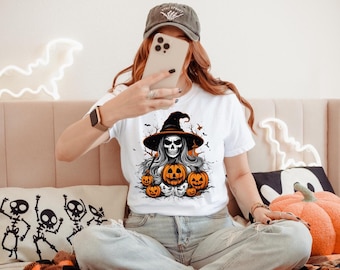 May include: A white short-sleeved t-shirt featuring a graphic print of a skeleton wearing a witch hat, surrounded by several orange jack-o'-lantern pumpkins, bats, and bare tree branches, set against a plain white background.