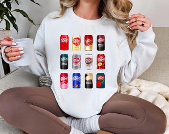 May include: White sweatshirt featuring a colorful graphic of various Dr. Pepper soda cans. The cans are in different colors and designs, including Zero Sugar and cherry flavors. The sweatshirt is a casual, comfortable style.