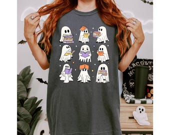 May include: Dark gray t-shirt featuring a grid of nine cartoon ghosts, each holding or surrounded by books. The ghosts are white with black eyes and are set against a dark gray background. The shirt is a casual style.