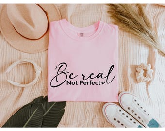 May include: A pale pink t-shirt with the words "Be real Not Perfect" in black script. The shirt is surrounded by a beige hat, white headband, scrunchie, sneakers, and dried plants.