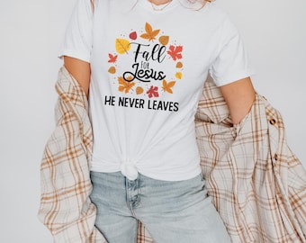 Puede incluir: Camiseta blanca con el texto "Fall for Jesus, He Never Leaves" rodeado de hojas de otoño. La camiseta se combina con vaqueros de lavado claro y una camisa a cuadros.