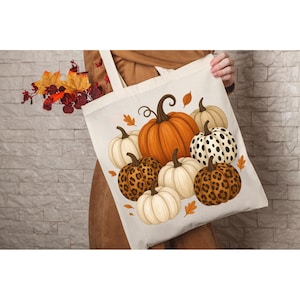 Könnte beinhalten: Beige Tragetasche mit einem Kürbismuster. Die Kürbisse sind orange, weiß und leopardenmuster, mit braunen Stielen und grünen Blättern. Die Tasche wird von einer Person in einem braunen Mantel gehalten. Im Hintergrund sind Herbstblätter und eine Backsteinmauer.