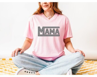 Op de afbeelding: Een roze T-shirt met een zwart geruit ontwerp en het woord "MAMA" in zwarte letters.
