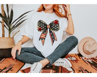 May include: White t-shirt with a large bow design in a patriotic style. The bow incorporates elements of the American flag, with stars and stripes in red, white, and blue. The shirt is worn with blue jeans and white trainers.