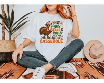 Puede incluir: Camiseta blanca con un pavo de dibujos animados con gafas de sol y zapatillas deportivas. La camiseta tiene el texto "TURKEY GRAVY BEANS & ROLLS LET ME SEE THAT CASSEROLE" en naranja, verde y marrón.
