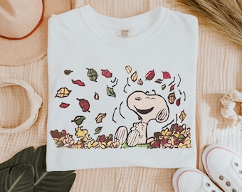 May include: A white t-shirt featuring a graphic print of the cartoon character Snoopy sitting in a pile of colorful autumn leaves. The design includes falling leaves in shades of red, yellow, green, and brown, with the Comfort Colors brand label visible.