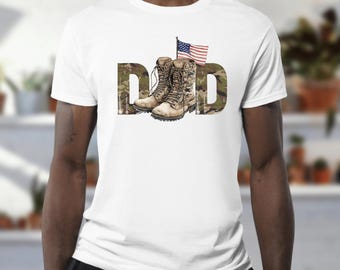 May include: White t-shirt with the word "DAD" in camouflage print. The "D"s frame a pair of combat boots and an American flag. The shirt is a crew neck and appears to be made of cotton.