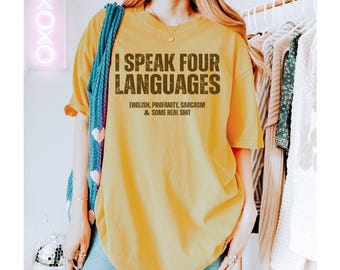以下が含まれることがあります： 前面に「私は4つの言語を話す：英語、悪口、皮肉、そしていくつかの本当のこと」という文字が印刷された黄色のTシャツです。