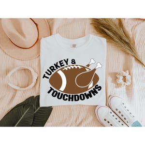May include: White t-shirt featuring a graphic combining a football and a turkey leg, with the words "TURKEY & TOUCHDOWNS" in a circular design. The shirt is on a light-coloured surface with a hat, scrunchie, and trainers.