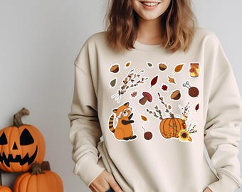 Pode incluir: Camisola bege com desenhos de autocolantes com tema de outono. Os autocolantes incluem um panda vermelho, abóboras, folhas, bolotas e um frasco de geleia. A camisola tem mangas compridas e gola redonda.