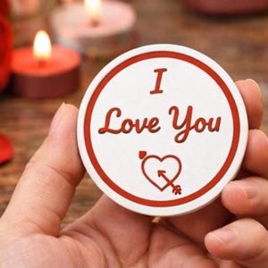 May include: A circular white and red token bearing the words "I Love You" and a heart with an arrow. The token is held in a hand, with red rose petals and lit candles in the background.