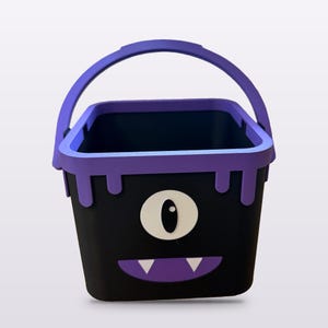 May include: A black square bucket with a purple handle and a purple rim featuring a dripping paint effect. The front displays a single large white eye with a black pupil and a purple mouth containing two small white triangular teeth.