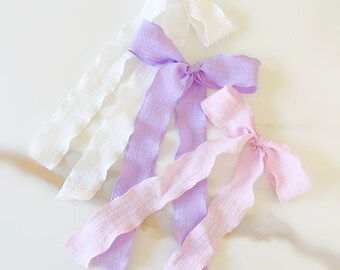 Può includere: Tre fiocchi per capelli in tessuto bianco, lavanda e rosa chiaro. I fiocchi sono realizzati in un tessuto strutturato con un bordo smerlato.