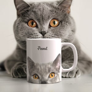 May include: A white ceramic mug with a gray cat face illustration and the word "Peanut" printed on the front. A gray cat is peeking over the top of the mug.