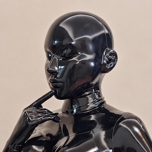 May include: A glossy black latex hood and matching bodysuit covering the head and neck. The material has a highly reflective, shiny finish. The headpiece features molded facial contours, including defined eyes, a nose, and lips, with one hand raised.