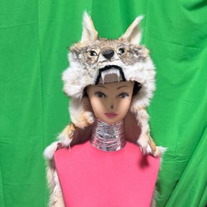 May include: A coyote fur hat featuring a realistic animal head with pointed ears, yellow eyes, and a black nose. The hat displays an open mouth with visible white teeth, and the fur is a mix of grey, brown, and white tones, covering the head.