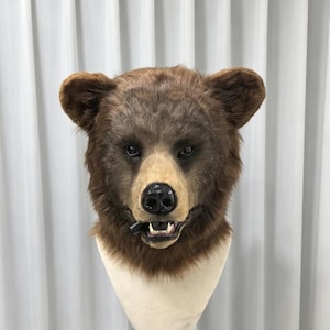 May include: A realistic brown bear head mask featuring dense, textured faux fur, rounded ears, and a light-colored muzzle. The mask includes a glossy black nose and an open mouth revealing white teeth, displayed on a neutral-colored mannequin head.