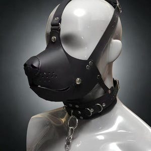 May include: A black leather animal-style mask with a snout, perforated nose detail, and adjustable straps. The mask is paired with a matching black leather collar featuring silver-toned metal hardware, including D-rings and a metal chain attachment.