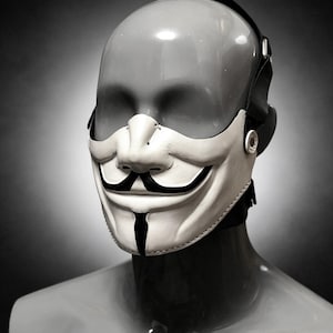 Pode incluir: Uma máscara de couro branco que cobre a metade inferior do rosto, apresentando um design estilizado de bigode e cavanhaque pretos. A máscara é fixada com tiras pretas ajustáveis e ferragens metálicas, exibida numa cabeça de manequim neutra.