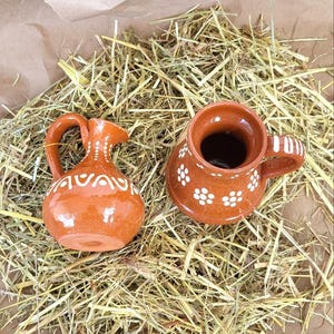 May include: Two small, brown ceramic pottery pieces with white floral designs. A pitcher and a mug are sitting on a bed of straw.