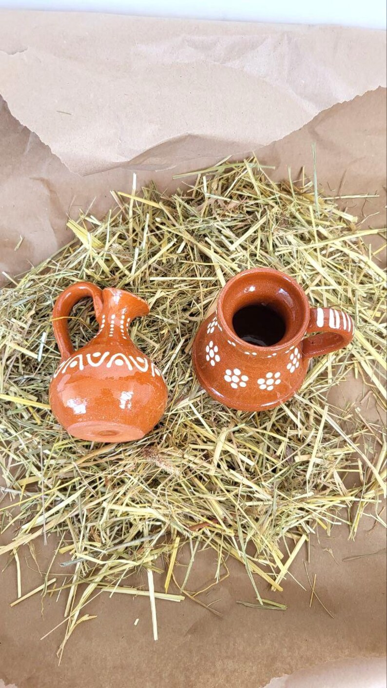 May include: Two small, brown ceramic pottery pieces with white floral designs. A pitcher and a mug are sitting on a bed of straw.