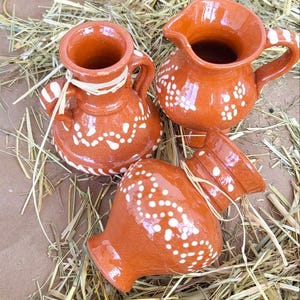 May include: Three small terracotta pottery jugs with white painted designs. The jugs are sitting on a bed of straw.