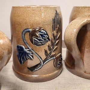 Op de afbeelding: Twee bruine keramische mokken met blauwe en bruine bloemenmotieven. De mokken hebben een handvat aan de zijkant.