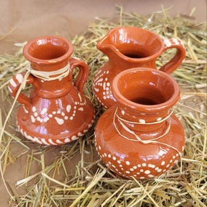 May include: Three small, terracotta pottery pitchers with white painted designs. The pitchers are sitting on a bed of straw.