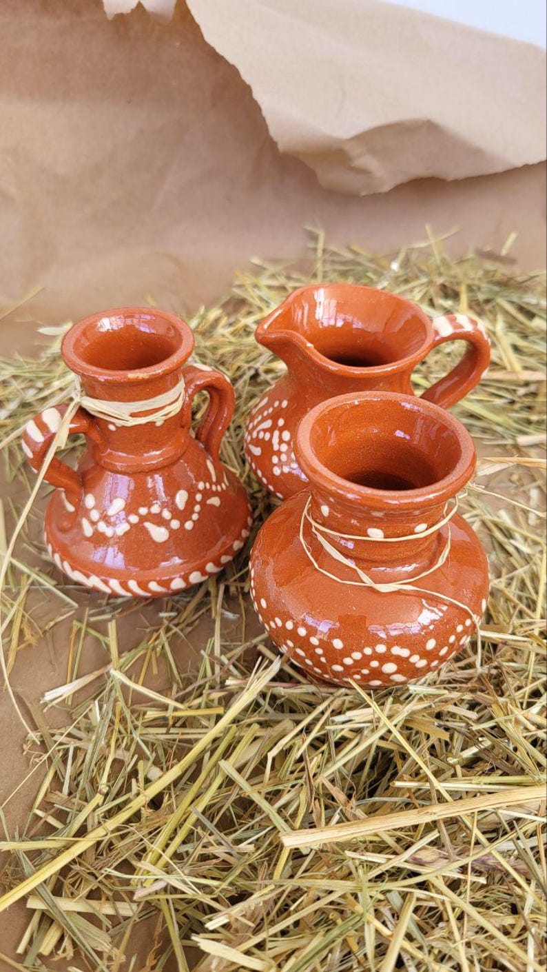 May include: Three small, terracotta pottery pitchers with white painted designs. The pitchers are sitting on a bed of straw.