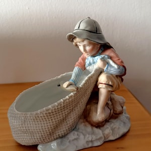 Könnte beinhalten: Porzellanfigur, die eine junge Person mit Fischerhut zeigt, die in einen geflochtenen Korb greift. Die Figur trägt ein rot-blau gestreiftes Hemd und beige Shorts. Der Korb steht auf einem hellblauen Sockel, der Wasser andeutet.