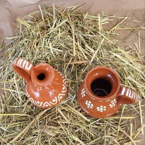 May include: Two small, brown ceramic jugs with white designs. The jugs are sitting on a bed of dried hay.
