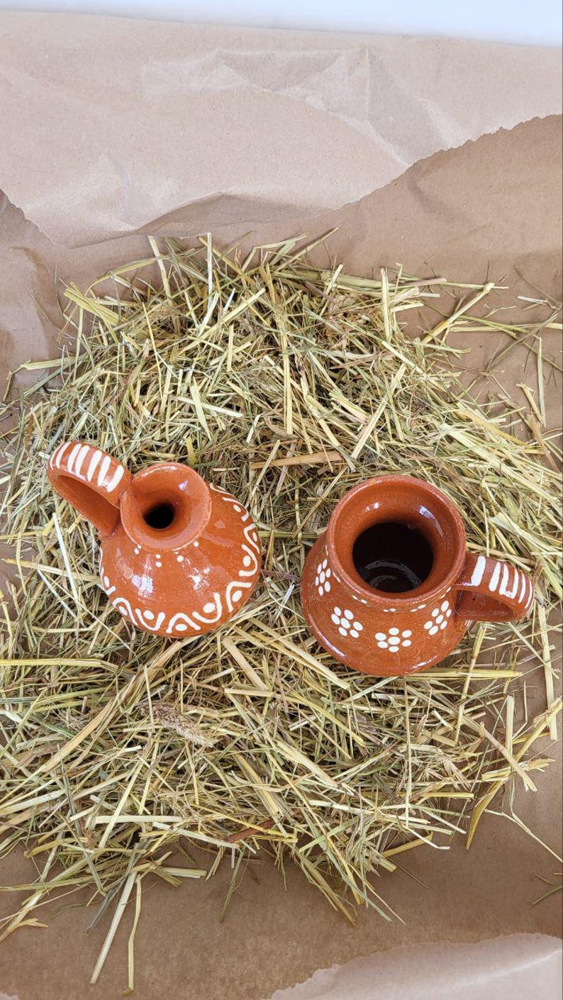 May include: Two small, brown ceramic jugs with white designs. The jugs are sitting on a bed of dried hay.