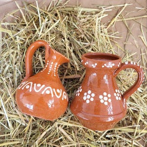 May include: Two small, brown ceramic pitchers with white designs. One pitcher has a handle and the other has a spout. Both pitchers are sitting on a bed of straw.