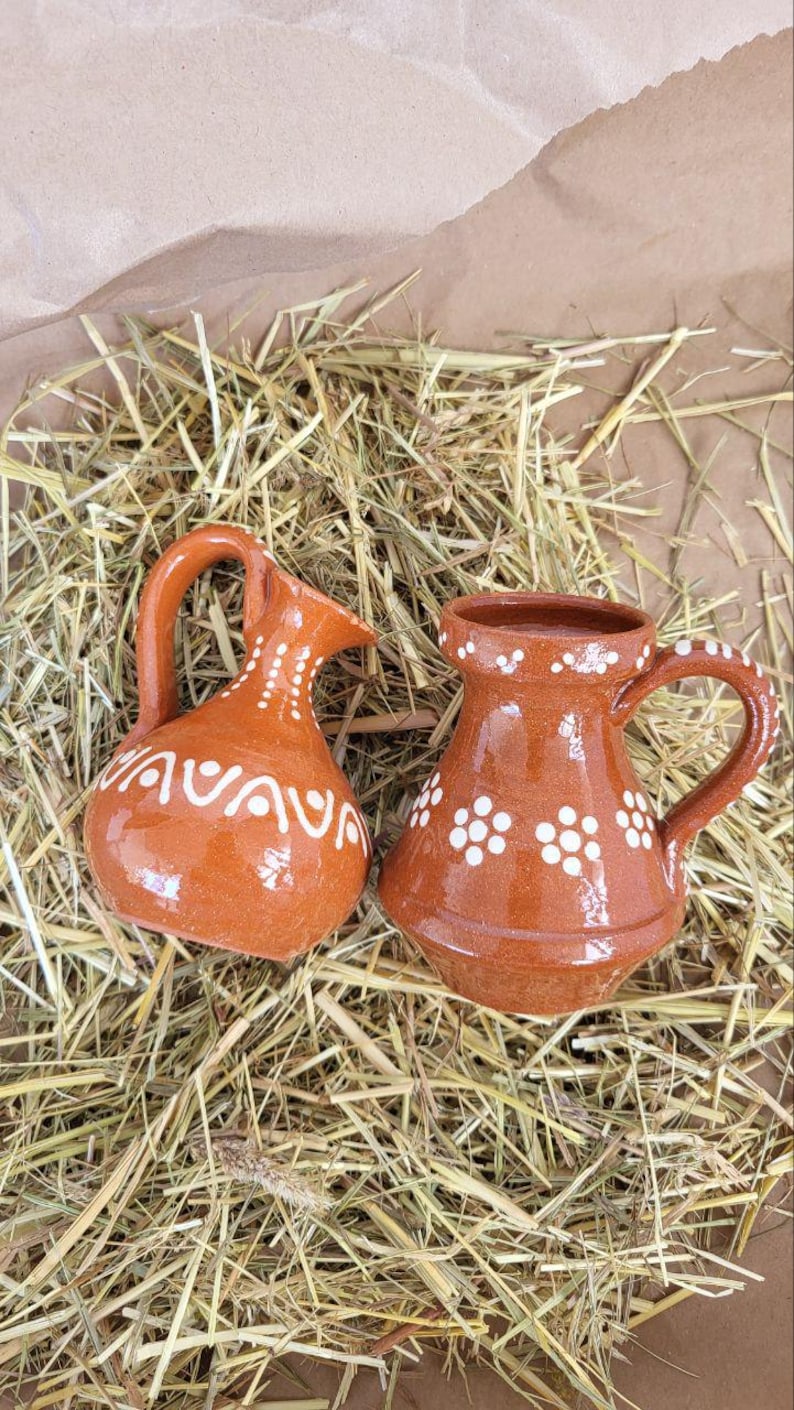 May include: Two small, brown ceramic pitchers with white designs. One pitcher has a handle and the other has a spout. Both pitchers are sitting on a bed of straw.