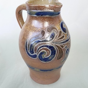 Peut inclure: Un pichet en céramique brun avec un motif floral bleu et brun. Le pichet a une anse et un bec.