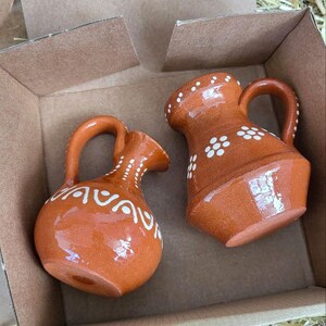 May include: Two small terracotta pitchers with white painted designs. The pitchers are in a cardboard box with straw packing material.
