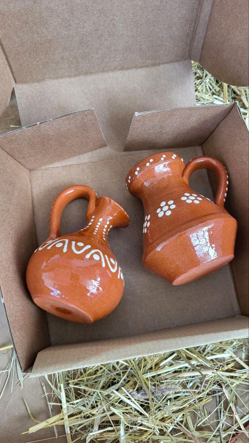 May include: Two small terracotta pitchers with white painted designs. The pitchers are in a cardboard box with straw packing material.