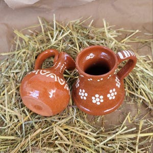 May include: Two small, brown ceramic pitchers with white floral designs. The pitchers are sitting on a bed of straw.