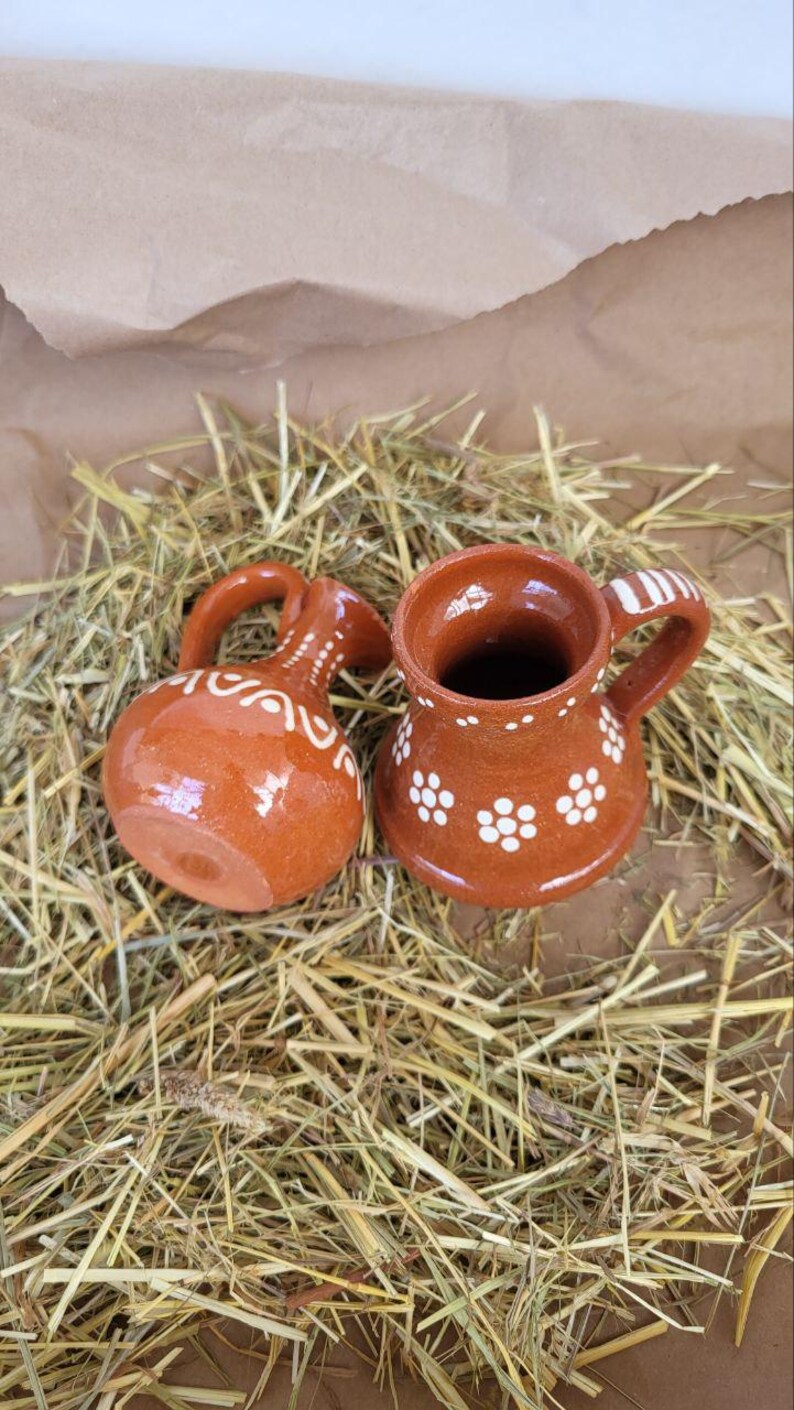 May include: Two small, brown ceramic pitchers with white floral designs. The pitchers are sitting on a bed of straw.