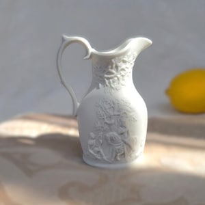Puede incluir: Una jarra de cerámica blanca con un diseño en relieve detallado de un árbol y figuras. La jarra tiene un asa y un pico.