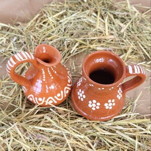 May include: Two small, brown clay pots with white painted designs. The pot on the left has a handle and a rounded body. The pot on the right has a handle and a wider, more cylindrical body.