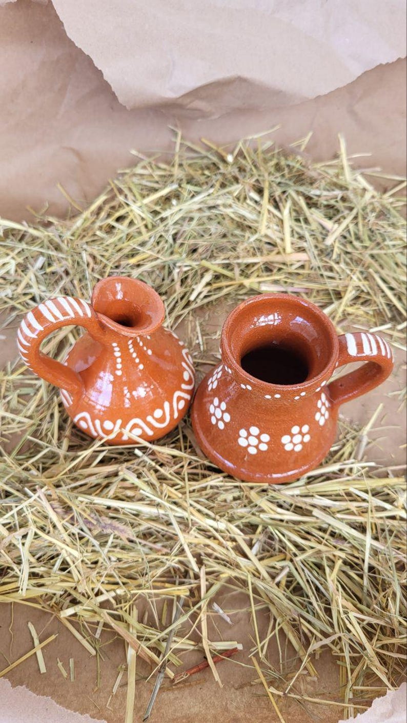 May include: Two small, brown clay pots with white painted designs. The pot on the left has a handle and a rounded body. The pot on the right has a handle and a wider, more cylindrical body.
