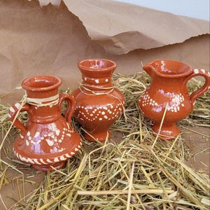 May include: Three small, terracotta pottery vases with white painted designs. The vases are sitting on a bed of straw.