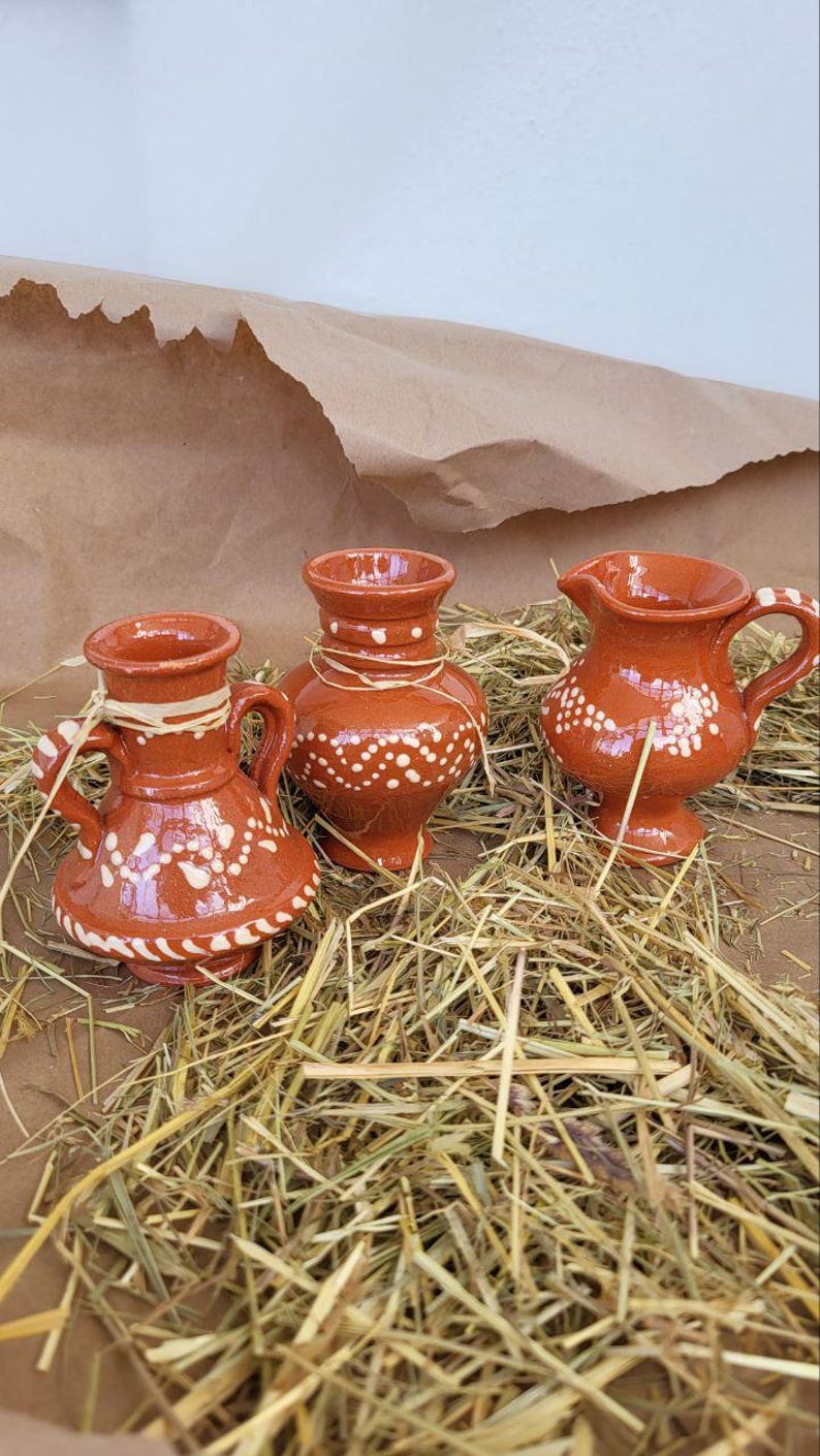May include: Three small, terracotta pottery vases with white painted designs. The vases are sitting on a bed of straw.