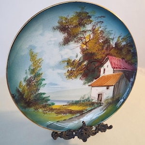 Op de afbeelding: Een rond keramiek bord met een geschilderd tafereel van een wit huis met een rood dak, bomen en een pad dat naar het huis leidt. Het bord heeft een gouden rand en staat op een decoratieve standaard.