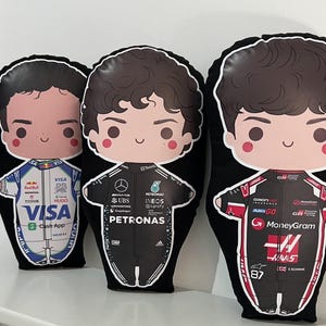 Puede incluir: Tres cojines de felpa negros con ilustraciones de dibujos animados de pilotos de Fórmula 1. Cada cojín presenta a un piloto con un traje de carreras con logotipos de patrocinadores. Los cojines tienen un borde blanco.