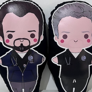 Pode incluir: Duas almofadas de pel&uacute;cia em forma de personagens de desenho animado. A almofada da esquerda mostra uma pessoa com cabelo escuro, barba, casaco azul-escuro com fecho e estetosc&oacute;pio. A da direita tem cabelo cinzento e veste uma t&uacute;nica preta com estetosc&oacute;pio.