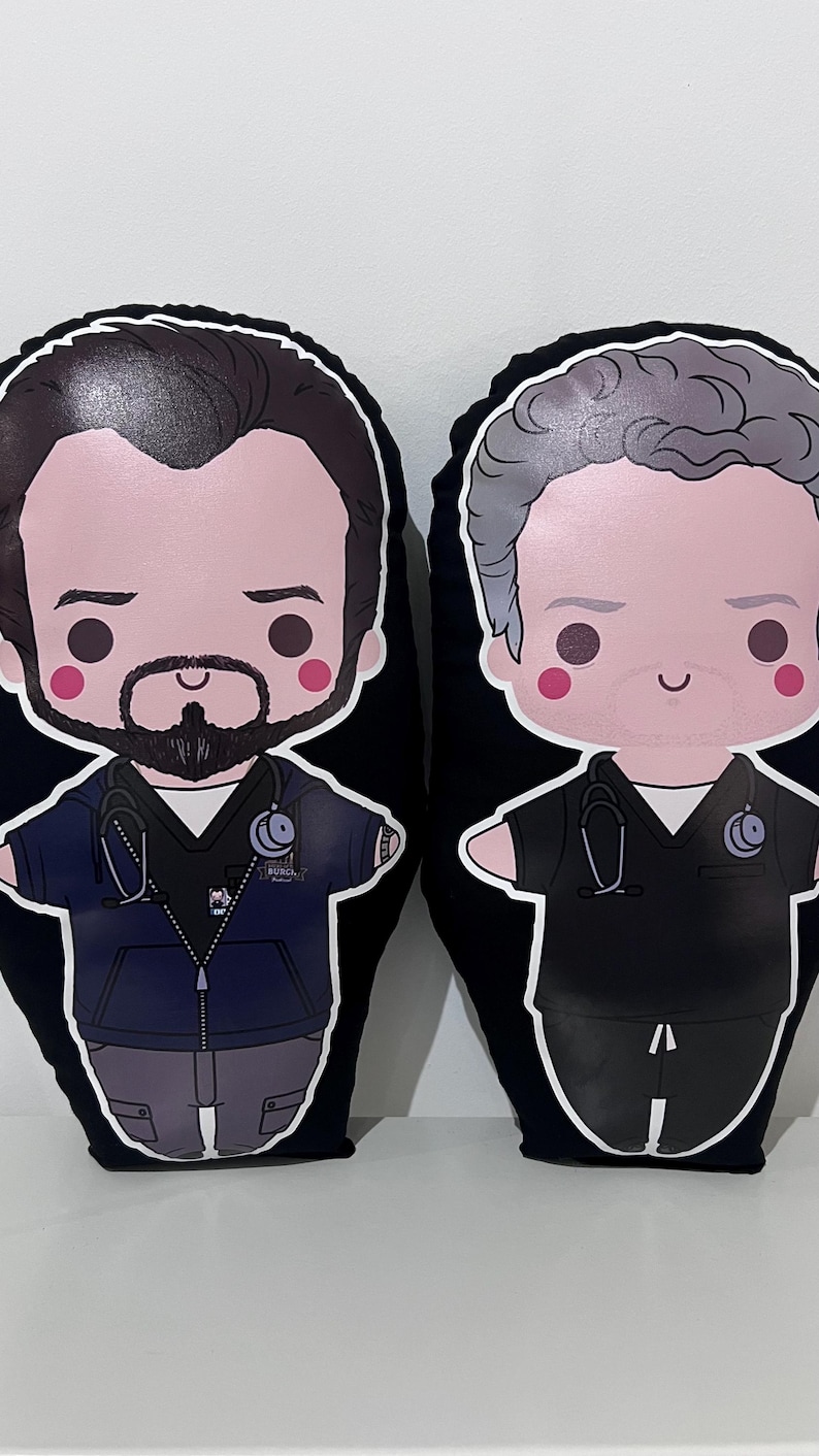 Pode incluir: Duas almofadas de pel&uacute;cia em forma de personagens de desenho animado. A almofada da esquerda mostra uma pessoa com cabelo escuro, barba, casaco azul-escuro com fecho e estetosc&oacute;pio. A da direita tem cabelo cinzento e veste uma t&uacute;nica preta com estetosc&oacute;pio.